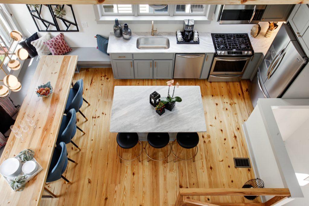 Aerial shot of an Heirloom kitchen in Savannah, GA. When we design vacation rentals for big groups, we make sure each home is special. Here, warm wood tones throughout immediately grab the eye, from the flooring, table, and ceiling beams. A dining table lines the left side of the photo, a breakfast bar island is in the center of the photo, and the sink and appliances go from the center-back of the image wrapping around the back along the right side.
