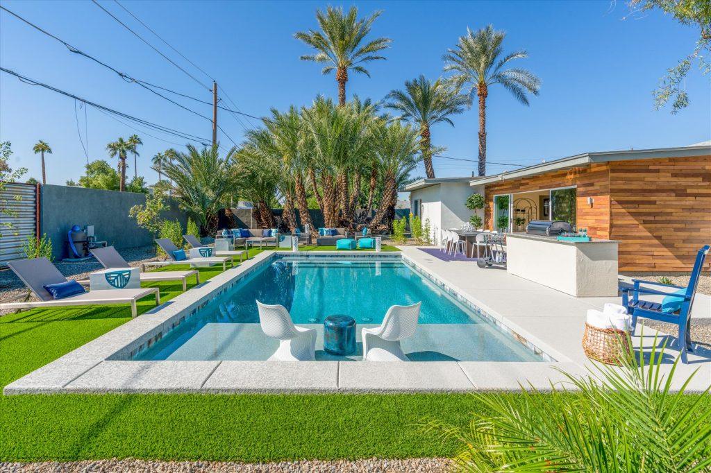 The sun shines down on a glittering, turquoise pool in the backyard of an Heirloom luxury home in Phoenix, AZ. Loungers line the pool as does a grill and outdoor kitchen, and palm trees rise in the background of the photo over the fire pit. The perfect vacation rental for big groups.