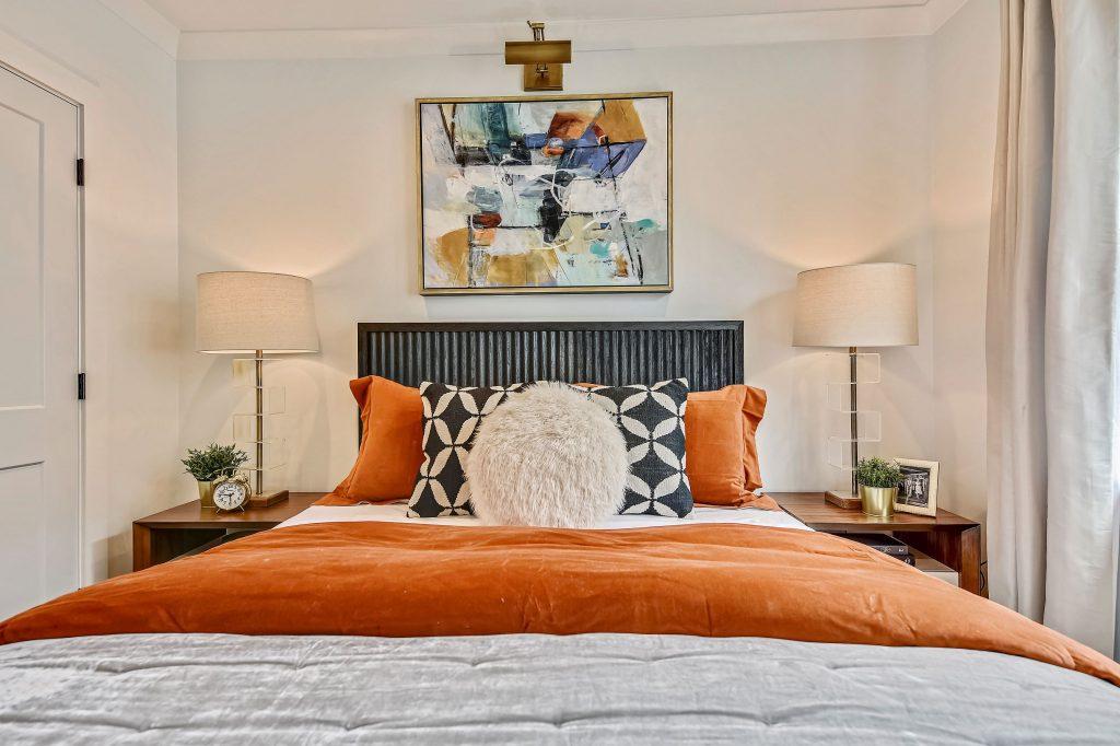 bed photographed head-on with orange duvet and white sheets. a poofy pillow is at the head of the bed. Two lamps line each side of the bed, and there is an abstract painting hung above it. the final example in our design interview.